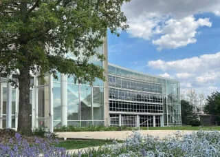 building with green trees, blue sky with white clouds, and garden with purple flowers surrounding it