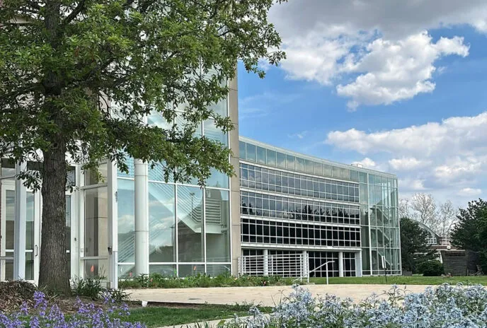 building with green trees, blue sky with white clouds, and garden with purple flowers surrounding it