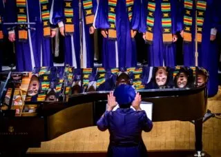 chorus wearing blue robes singing and reflected in black piano