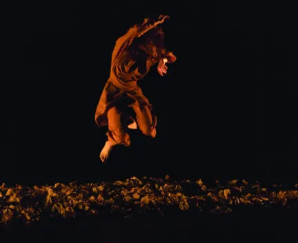 dancer leaps in the air in orange lighting, background is dark, and floor is littered with paper-like confetti