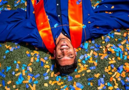 Male student lying on their back in the grass in graduation garb with orange and blue confetti around them