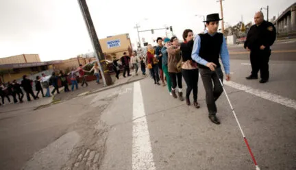 a person uses a white cane on a road while a line of people hold on one by one with their eyes closed