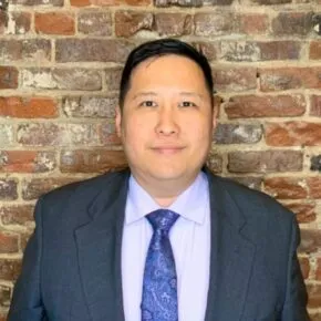 person wearing a grey suit with white shirt and purple paisley tie stands in front of brick wall