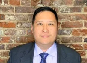person wearing a grey suit with white shirt and purple paisley tie stands in front of brick wall
