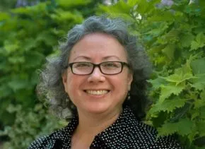 person with short gray hair wearing glasses and dark shirt with white polka dots smiles widely and stands in front bright green bush