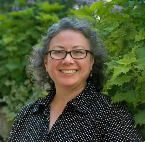 person with short gray hair wearing glasses and dark shirt with white polka dots smiles widely and stands in front bright green bush