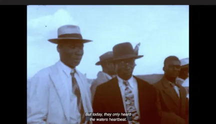 A group of Black men are standing next to each other. They are dressed in suits, ties and hats. Above them is a wide expanse of sky. The colors in this still image from Handsworth Songs are very muted, almost black and white. The captions at the bottom of the screen read "But today, they only heard the waters heartbeat."