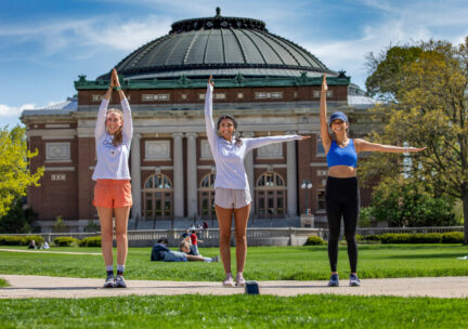 three students on the central green grassy quad hold up their arms to make 