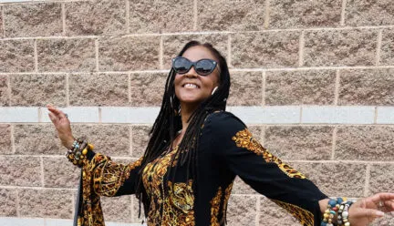 nadirah wearing black and gold and dark sunglasses with arms out and smiling, light stone wall in the background