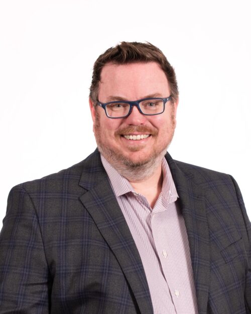 man with brown hair, facial hair, glasses, in a dark gray suit with lavender shit