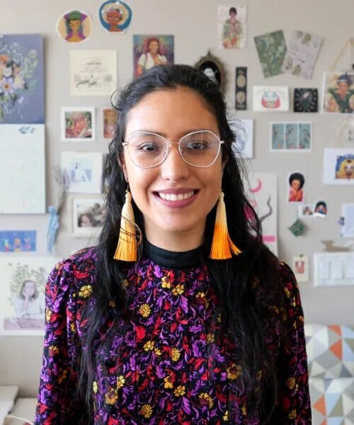 person wearing long tassle orange earrings, light-rimmed glasses, and a colorful shirt, smiling