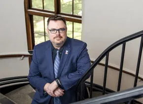 man with brown hair, facial hair, glasses, in a dark gray suit with lavender shit