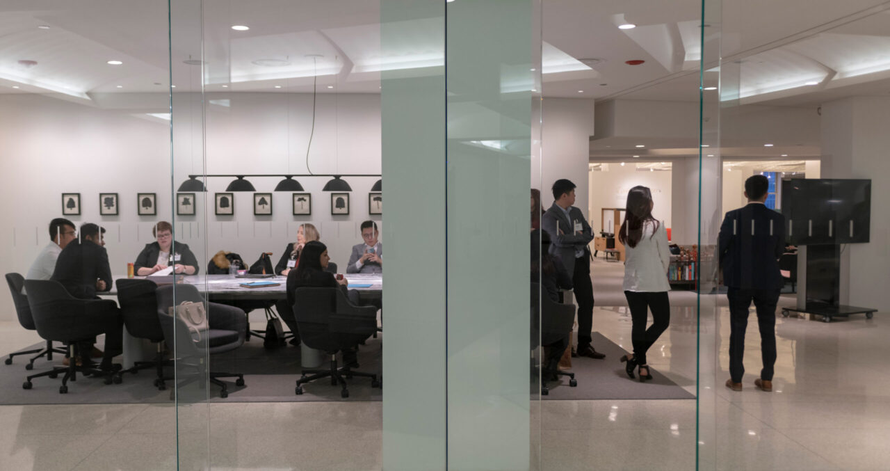 people in business attire sit at a conference table behind a glass partition