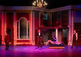 stage set that looks like a luxurious early 20th century home drenched in pink and purple lighting with actors onstage