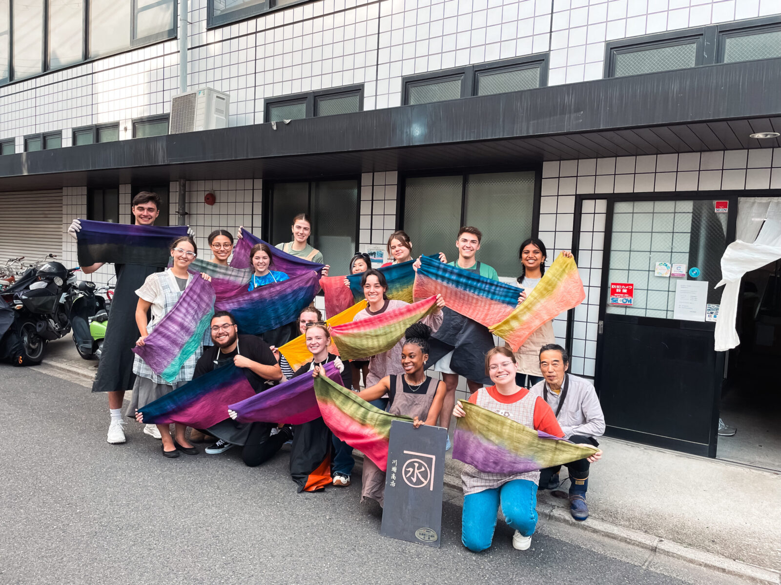 15 people holding up colorful fabric on small street