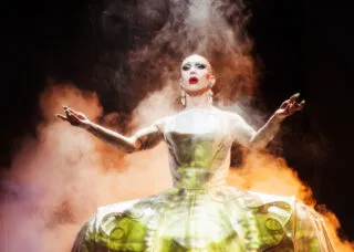 drag queen in large gold dress on stage with arms extended