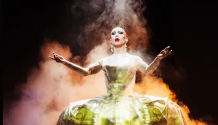 drag queen in large gold dress on stage with arms extended