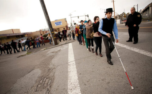 a person uses a white cane on a road while a line of people hold on one by one with their eyes closed