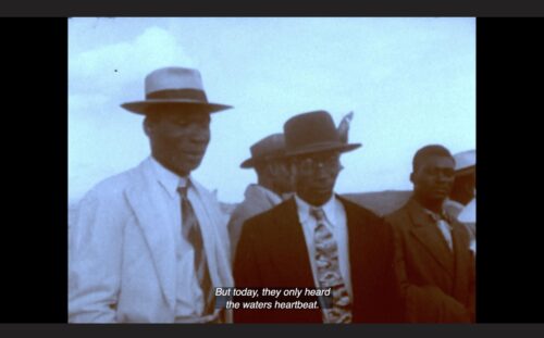 A group of Black men are standing next to each other. They are dressed in suits, ties and hats. Above them is a wide expanse of sky. The colors in this still image from Handsworth Songs are very muted, almost black and white. The captions at the bottom of the screen read 