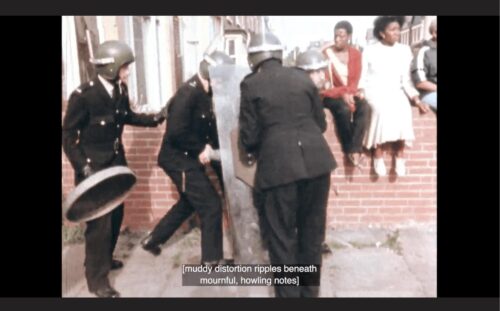 A still image from the film Handsworth Songs. A group of three white policemen equipped with shields and helmets surround someone. Behind them on a brick wall three Black people are sitting. The captions at the bottom of the image read “[muddy distortion ripples beneath mournful, howling notes]”.