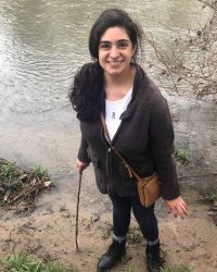 person stands by a body of water holding a wooden stick and wearing a gray sweater and light brown bag