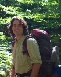 person stands by trees wearing light green shirt and large red and black backpack with gear