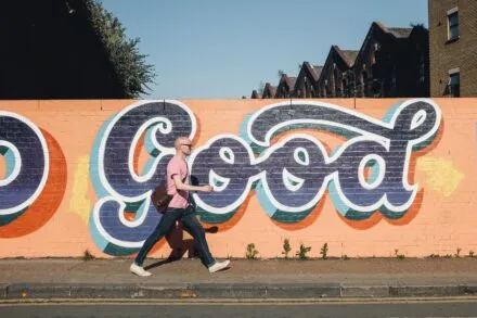 A man with backpack walking on the sidewalk in front of a brick wall with "good" painted in cursive on it