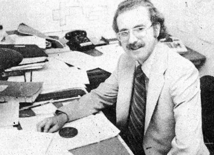 Black and white photo of Doug Houston sitting at office desk surrounded by papers and maps on the wall.