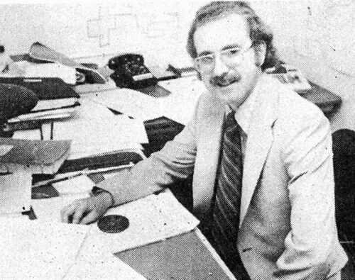 Black and white photo of Doug Houston sitting at office desk surrounded by papers and maps on the wall.