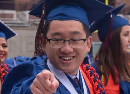 Photo of Zishen Ye in blue cap and gown with orange cords pointing to camera
