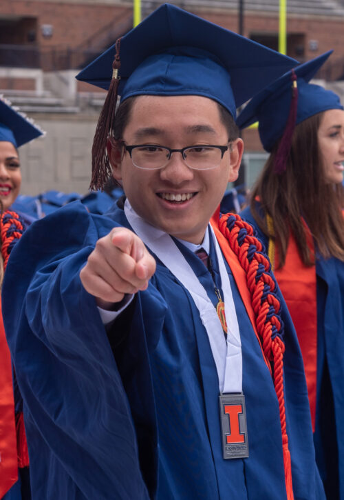 Photo of Zishen Ye in blue cap and gown with orange cords pointing to camera