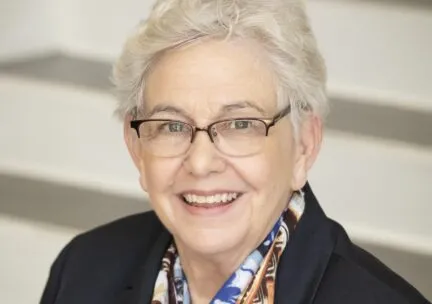 caucasian wearing blue blazer, colorful blouse, white permed hair, round glasses, smiling at camera