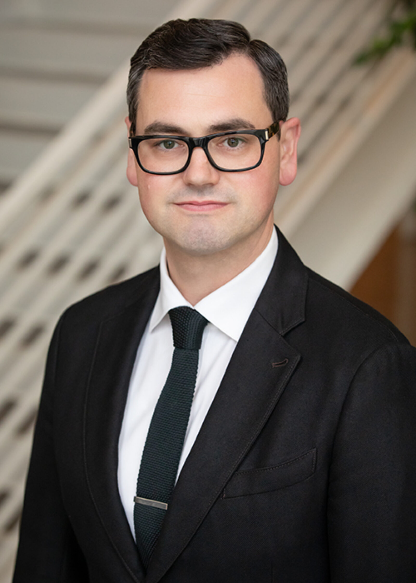 man smiling at camera wearing black rimmed glasses black suit black tie white button down shirt