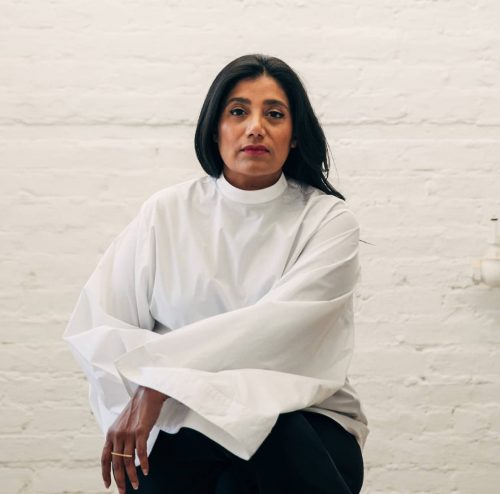 Suchi Reddy seated against a white brick background 