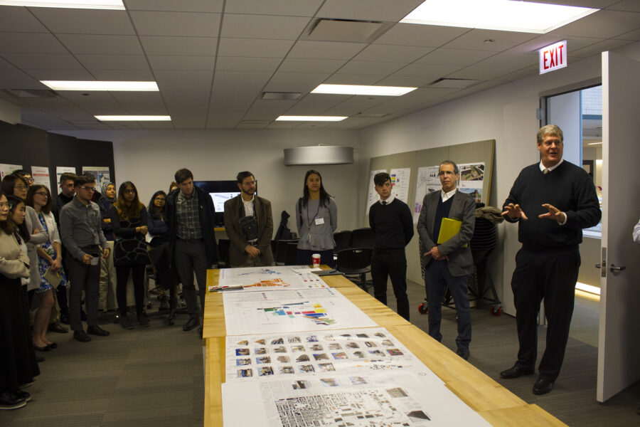 students and faculty standing around a long table with student work on it 