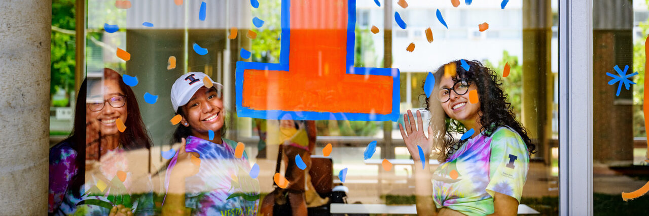 three girls looking outside of a window, smiling at the camera, orange I on the window with window confetti stickers 