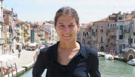 woman standing on bridge, water behind her, houses on the sides, black shirt orangish skirt