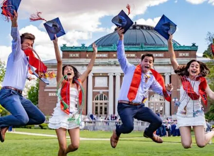 four students jumping, excited to have graduated, smiling, holding their graduation hats in their hands, in front of foellinger auditorium in the main quad