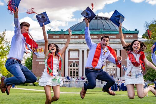 four students jumping, excited to have graduated, smiling, holding their graduation hats in their hands, in front of foellinger auditorium in the main quad