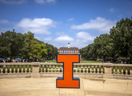Orange I sitting on foellinger autitorium balcony in front of the illini union, blue sky, white clouds, green trees 