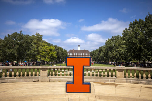 Orange I sitting on foellinger autitorium balcony in front of the illini union, blue sky, white clouds, green trees 