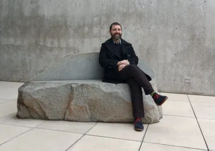 a man smiling with his legs crossed, dressed all in black sitting on a rock with a graphite background