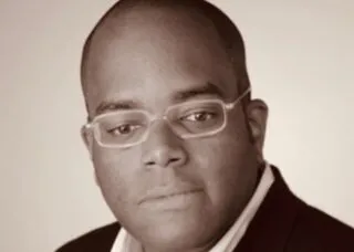 man staring under camera, wears glasses, brown complexion, no hair, black suit white button down undershirt