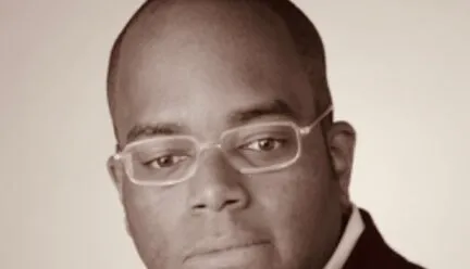 man staring under camera, wears glasses, brown complexion, no hair, black suit white button down undershirt