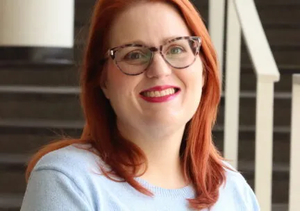 woman smiling at camera, red hair, glasses, light blue shirt