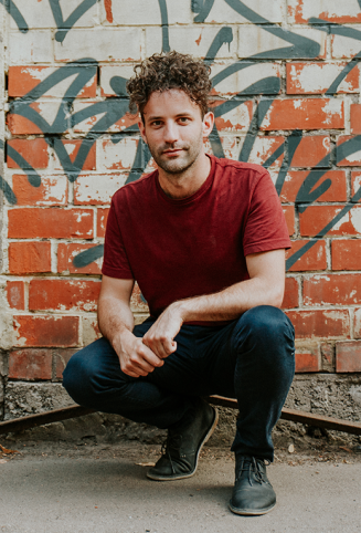 man looking at camera with graffiti behind him squatting with jeans and red shirt