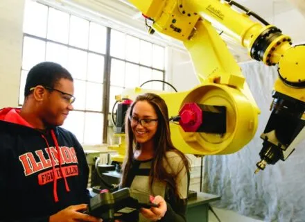 two students smiling next to a robotic arm