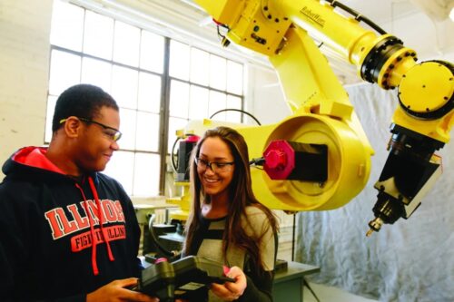 two students smiling next to a robotic arm