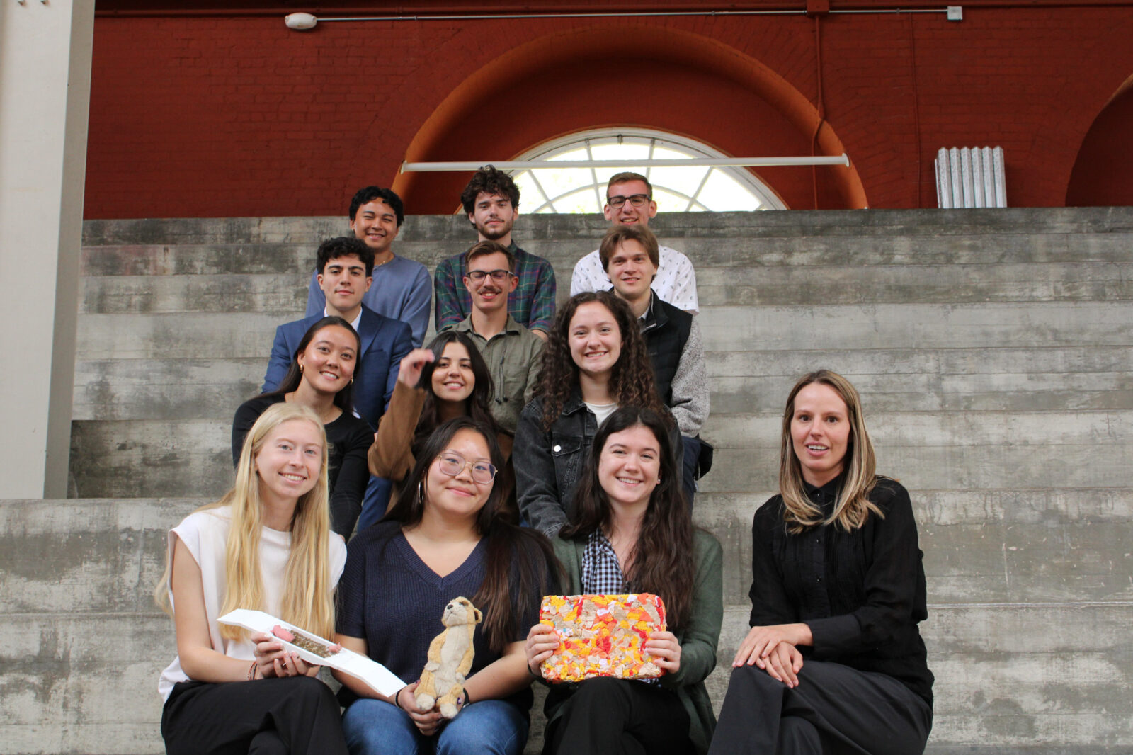 12 students with their professor sitting on steps at the stock pavilion holding their models