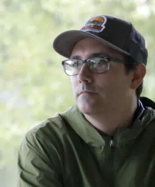 Portrait of Ryan Griffis in an outdoor setting, wearing a baseball hat and green rain jacket.